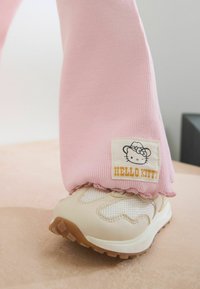 Różowe spodnie z prążkowanego materiału z falbaniastym dołem mają metkę "Hello Kitty". Beżowe trampki z siateczkowymi i gumowymi podeszwami dopełniają strój.