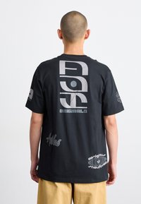 Svart bomulls-t-shirt med en stor grå grafik på baksidan, med texten "ASG Originals" och flera mindre logotyper på sidorna.