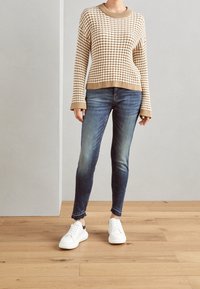 Suéter de punto a cuadros en beige y blanco con mangas anchas, combinado con jeans ajustados azules y zapatillas blancas con detalles en negro.
