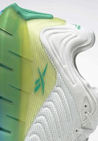 Basket en blanc avec une tige en maille texturée, semelle translucide verte et jaune, et détail de marque turquoise. Dotée d'un col rembourré.