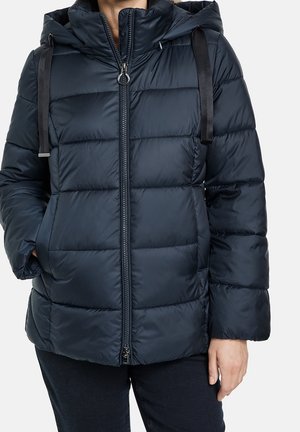 Doudoune matelassée bleu marine avec capuche, cordons noirs, fermeture éclair à l'avant, mains dans les poches, portée avec un pantalon foncé.