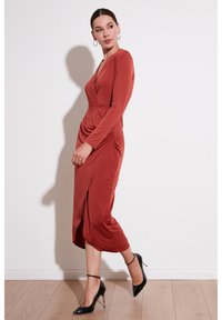 Robe de couleur rouille à manches longues avec un décolleté en V, un devant drapé et une fente sur le côté. Portée avec des chaussures à talons hauts noires et des boucles d'oreilles en créole.