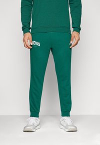 Sötétzöld melegítőnadrág, elasztikus mandzsettákkal, fehér "ADIDAS" logóval a bal combon. Az anyag puhanak és rugalmasnak tűnik.