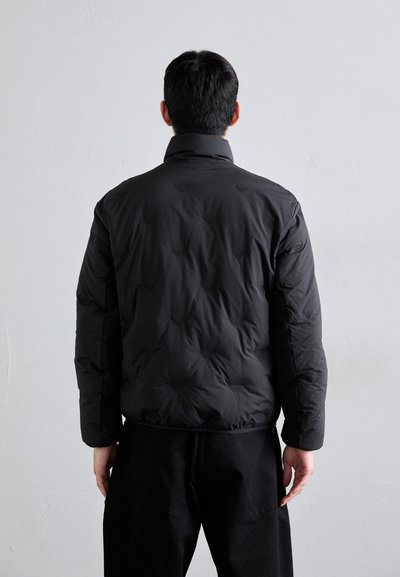 Casaco puffer preto com gola alta, apresentando uma textura acolchoada, mangas longas e um design aerodinâmico, usado sobre calças pretas.