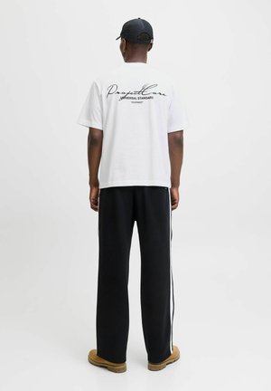 White short-sleeve t-shirt with black cursive text on the back, paired with black pants featuring white side stripes and brown footwear.