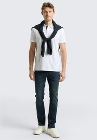 Biała koszulka polo z czarnym kołnierzykiem, granatowy sweter związany wokół ramion, ciemne jeansy i białe trampki, stojąca na tle jasnoszarego tła.