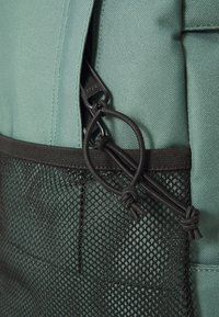Rucsac din material verde cu o buzunare din plasă neagră texturată. Dispune de un fermoar și un cordon negre, având un accent de hardware cu bucle pentru asigurarea obiectelor.