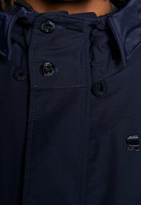 Marinblå jacka med huva och svarta knappar, justerbar dragsko och diskret logotypdetalj. Har slät, texturerad tyg och sömdetaljer.