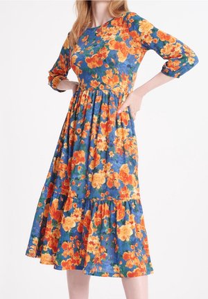 Mujer con vestido midi azul con estampado floral en naranja y amarillo, mangas tres cuartos y cintura fruncida, de pie con las manos en las caderas.