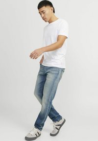 Camiseta de algodón blanca, jeans de mezclilla azul con un acabado desgastado, zapatillas grises con acentos negros, en pose de pie con un pie hacia adelante.