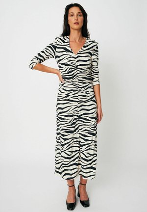 Kobieta stojąca z ręką na biodrze, ubrana w midi sukienkę w czarno-biały wzór zebry oraz czarne szpilki z paskiem na kostce, na jednolitym tle.