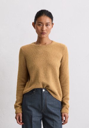 Senfgelbes, strukturiertes Strickpullover mit langen Ärmeln, rundem Ausschnitt und gerippten Bündchen, kombiniert mit dunklen Jeanshosen.