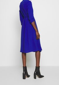 Långärmad, blå klänning med midjebälte, utsvängd nederdel och en mjuk textur, kombinerad med svarta ankelskor och klack.