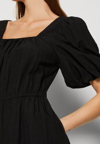 Femme portant une robe noire avec manches courtes bouffantes, encolure carrée et taille élastiquée, visible du cou à la taille.