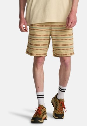 Dolna część ciała osoby noszącej beżowe szorty w brązowe i zielone paski, białe skarpetki z czarnymi paskami oraz brązowe buty turystyczne z pomarańczowymi sznurowadłami.