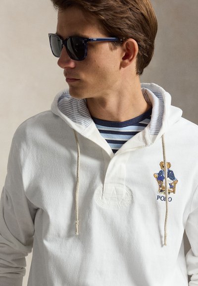 Jeune homme portant des lunettes de soleil foncées et un sweat à capuche blanc avec une broderie d'ours sur une chemise à rayures bleues, regardant vers la gauche.