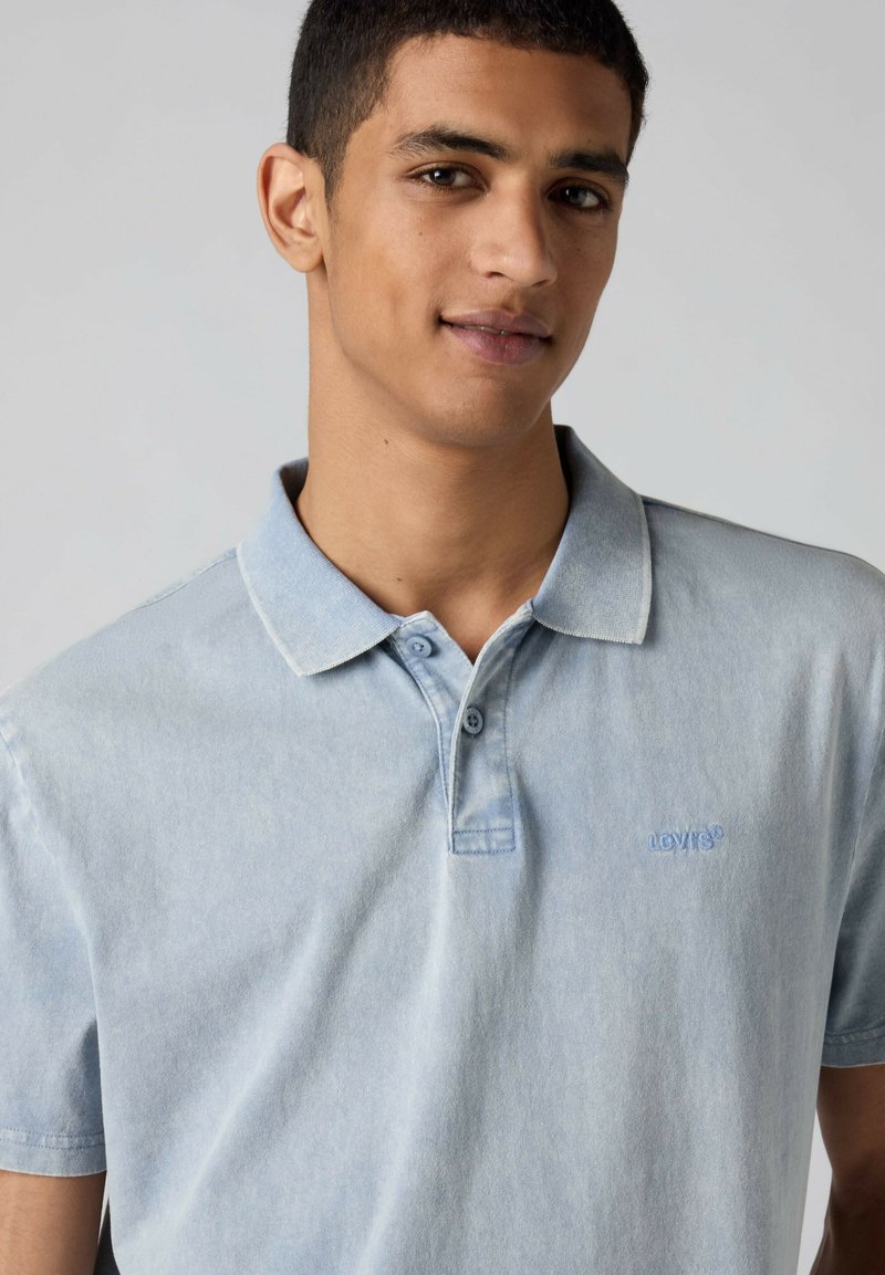 Young man wearing a light blue Levi's polo shirt, looking slightly to the side with a neutral expression on a plain background.