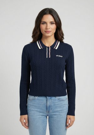 Mujer con suéter polo de manga larga de punto trenzado azul marino con el logo "GUESS" y jeans azul claro, de pie mirando hacia adelante.