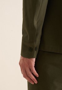 Manche de chemise vert olive avec détail de poignet à double bouton. Tissu lisse et structuré avec une coupe ajustée, mettant en valeur une main fine.