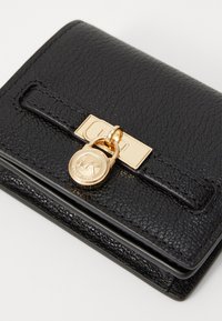 Fekete, textúrált bőrtárca arany Michael Kors kerek charm-mal és téglalap alakú csattal, fehér háttéren.