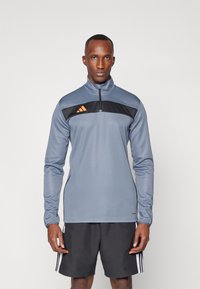 Sudadera atlética gris con cuello alto, cierre de media cremallera y panel en el pecho negro con tres rayas naranjas. Hecha de tejido suave.