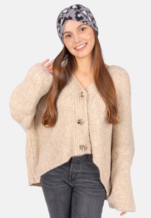 Beige Strickjacke mit V-Ausschnitt, großen Knöpfen und überschnittenen Schultern. Kombiniert mit dunkelgrauen Jeans und einem grauen gemusterten Stirnband.