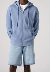 Felpa con zip di colore blu chiaro, con tasche frontali e cappuccio regolabile tramite lacci, abbinata a pantaloni corti in denim chiaro. Presenta una piccola etichetta rossa con il logo Levi's.