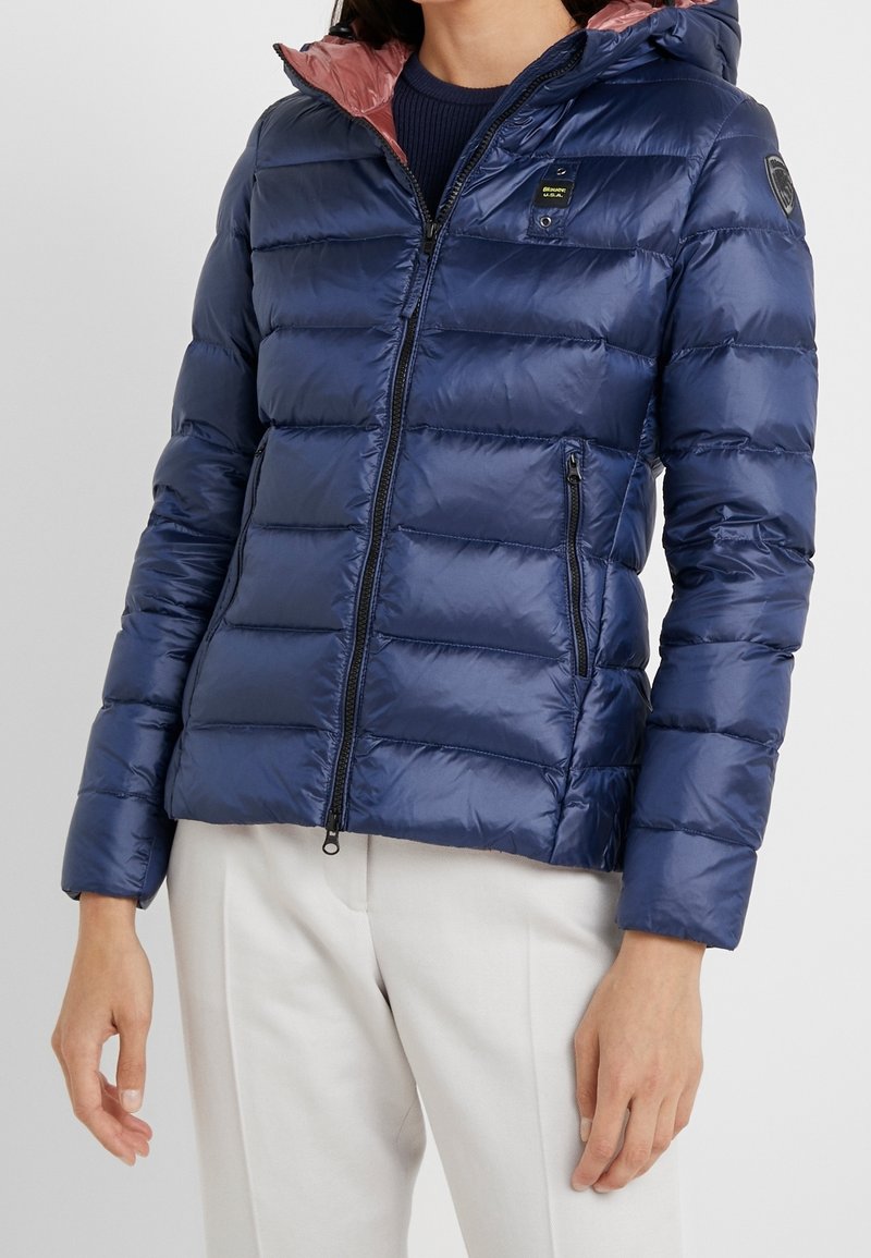 Marineblaue Puffjacke mit gestepptem Design, Kapuze und hellrosa Innenfutter. Sie verfügt über Reißverschlusstaschen und ein Logo-Emblem am Ärmel.