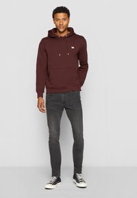 Bordeauxfarbener Hoodie aus Baumwollmischung; mit einer frontalen Kängurutasche, Kapuze mit Kordelzug und gesticktem Logo auf der Brust. Kombiniert mit grauen Jeans.