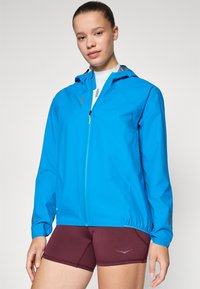 Young woman wearing a blue hooded jacket over a white top and maroon athletic shorts, standing against a plain background.