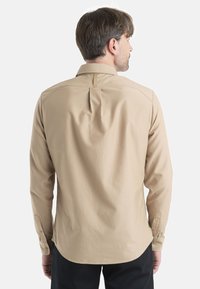 Camicia beige a maniche lunghe con yoke posteriore, rifiniture curate e polsini con bottoni, caratterizzata da un dettaglio a plissé sul retro. Design con colletto piatto.