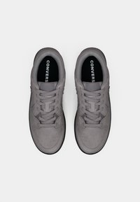 Grå semsket sneakers med flate lisser, perforert tå og svart gummisåle. "CONVERSE"-logo synlig på innersåler og side.