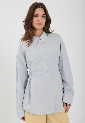 Svetlomodrá košeľa s vertikálnymi pásikmi v žltej farbe. Má klasický golier a dlhé rukávy s voľným strihom. Mäkký materiál.