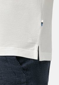 Hellgraues Polohemd, strukturierter Stoff, seitliche Schlitze am Saum, kleiner Farbakzent in Blau und Weiß an der Seitennaht.