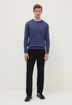 Hombre de pie vistiendo un jersey azul con cuello redondo, camiseta blanca, pantalones negros y zapatos de vestir negros contra un fondo liso.