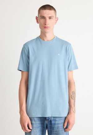 Light blue cotton T-shirt with a rounded crew neck, short sleeves, and a small CK logo on the chest. Worn with light denim jeans.