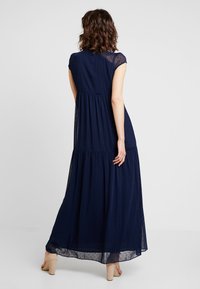 Žena stojící zády k fotoaparátu ve tmavě modrých šatech s krátkým rukávem a délce po kotníky, vyrobených z průhledného materiálu, doplněná o tělové lodičky.