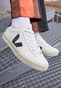 Weiße Ledersneaker mit einer strukturierten Sohle, schwarzen Seitenelementen und weißen Schnürsenkeln. Tragen sie mit hellen Socken und bunten Hosen auf einer Metalltreppe.