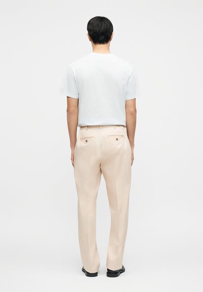 Ροζ ανοιχτό ροζ παντελόνι με ίσια γραμμή, δύο πίσω τσέπες με χρυσά κουμπιά, φορεμένο με λευκό t-shirt, φαίνεται από πίσω.