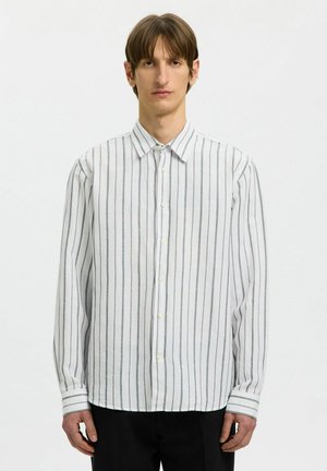 Jeune homme portant une chemise blanche à boutons avec des rayures noires verticales, debout devant un fond blanc uni.