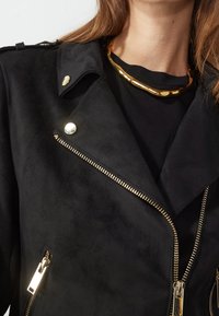 Zwarte suède jas met metallic gouden ritsen en drukknopen, met een reverskraag en een gladde textuur. Geaccentueerd met een gouden halsketting.
