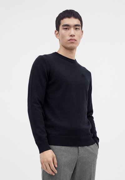 Homme portant un pull noir à manches longues et un pantalon gris, debout avec une main dans la poche devant un fond clair uni.