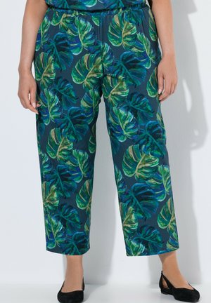 Frau trägt blau-grün gemusterte Hosen mit weitem Bein und schwarzer Ballerinas, steht vor einer weißen Wand.