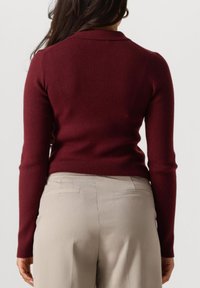 Vrouw in een aansluitend bordeauxrood geribd longsleeve shirt en beige op maat gemaakte broek, van achteren zichtbaar tegen een effen achtergrond.