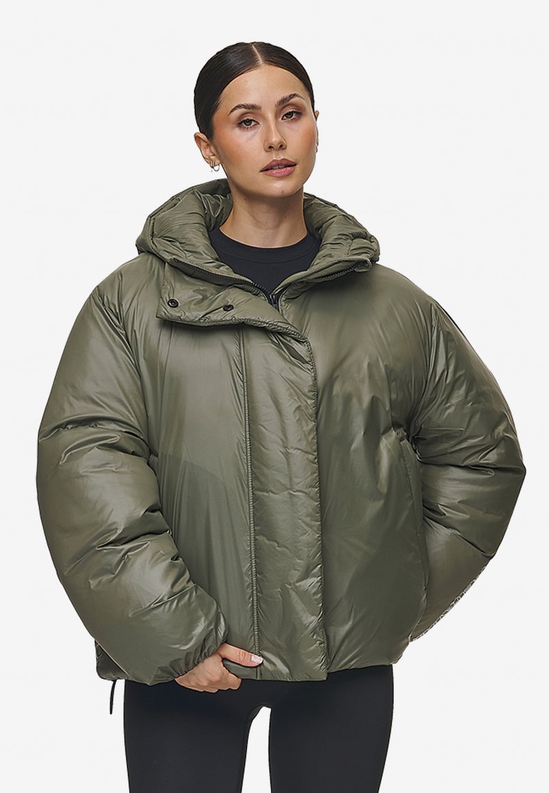 Olijfgroene pufferjas met een capuchon, voorzien van een ritsluiting, gewatteerde textuur en een oversized pasvorm. Middenlang met aanpassingsmogelijkheden.