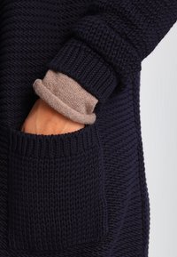 Marinblå stickad tröja med ett strukturerat mönster, försedd med en vikt beige manschett och en frontficka. Tyget verkar tjockt och varmt.