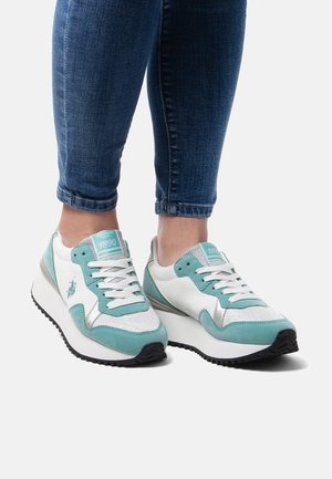 Persona che indossa sneakers con lacci turchese e bianche con suola platform e jeans blu tagliati sopra le caviglie, in piedi su sfondo bianco.