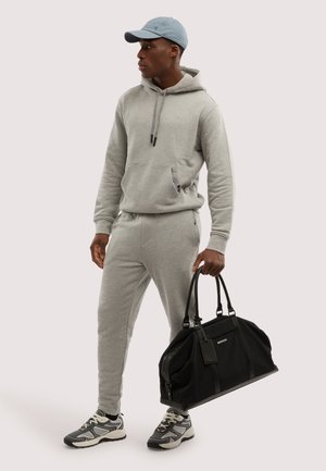 Homme en sweat à capuche gris et pantalon de survêtement, casquette bleue, baskets, tenant un grand sac de sport noir avec deux poignées.