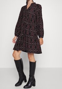 Svart klänning med röd och vit diamantmönster, lång puffärm och utsvängd nederkant, kombinerad med knähöga svarta klackstövlar.