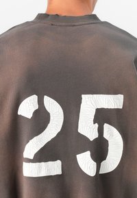 Sweatshirt cinza escuro com grandes números brancos "25" rachados nas costas. O tecido tem uma textura suave com ligeiro desgaste na gola.